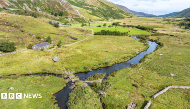 A green valley in Eryri national park. It has steep sides and is dotted with trees, a winding stream and a small stone cottage