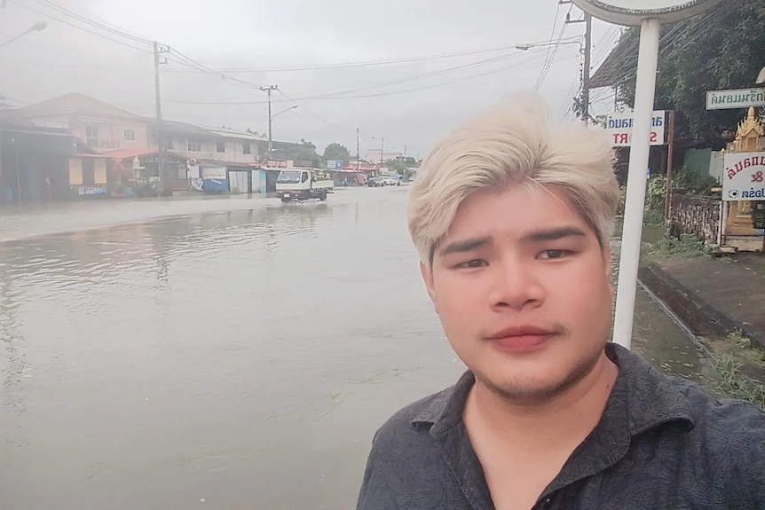 A Thai man with white blond hair stands in front of a completely flooded street.