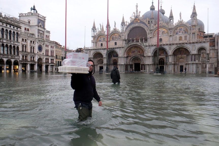 A man carrying packages in thigh-deep floodwaters.