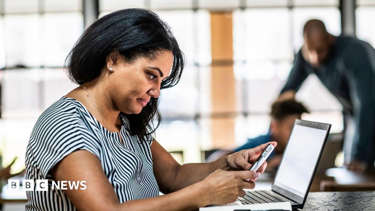 Woman looking at a phone sits at a desk with a laptop in front of her and a man and boy in the background.