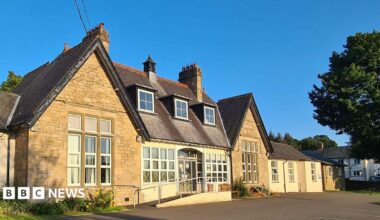 Alston Medical Practice is a white bungalow building with a black slate roof and has four sash windows. There is a small sign by the front door and a green bush and front yard. It is a sunny day with bright blue sky.