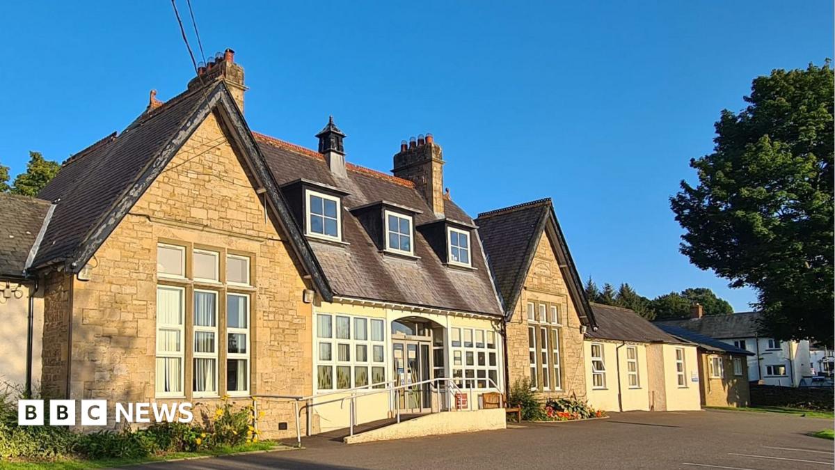 Alston Medical Practice is a white bungalow building with a black slate roof and has four sash windows. There is a small sign by the front door and a green bush and front yard. It is a sunny day with bright blue sky.