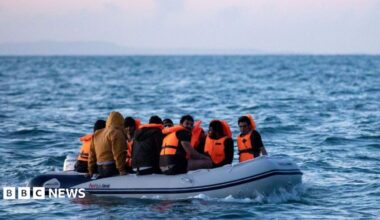 Migrants on a small inflatable boat attempt to cross the English Channel near the Dover Strait, the world's busiest shipping lane. The people on board the dinghy are wearing orange life jackets. The waves are splashing around the vessel. One person without a life jacket is wearing a brown jacket with a hood.