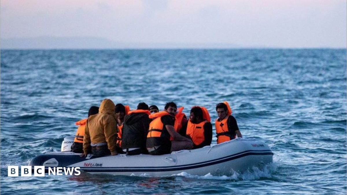 Migrants on a small inflatable boat attempt to cross the English Channel near the Dover Strait, the world's busiest shipping lane. The people on board the dinghy are wearing orange life jackets. The waves are splashing around the vessel. One person without a life jacket is wearing a brown jacket with a hood.
