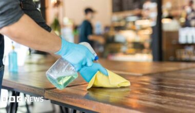 Person wearing blue disposable gloves sprays cleaning solution onto a wooden table and wipes the surface with a yellow cloth inside a café or restaurant, with the background softly blurred.