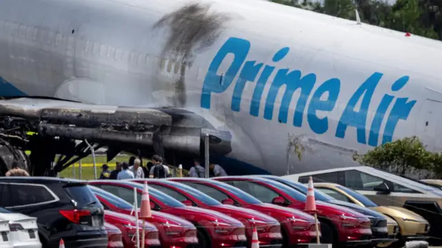 Scene of a crash where an Amazon Prime Air cargo plane overran the runway at Miami International Airport. A line of parked cars can be seen in front of the plane which is nose down and has burn marks on it