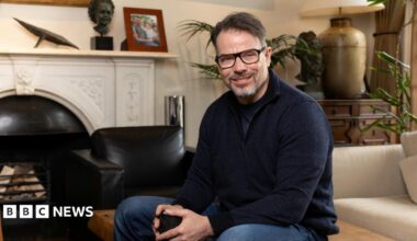 Chris Rokos sitting in a room wearing a dark blue jumper and blue jeans. In the background there is a fireplace, sofa and chest of drawers.