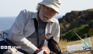 A man rests on his side on the ground as a small puffin bird sits in front of him. There is a cup of water with a paintbrush in it next to them. The man wears a cream bucket hat, white chequered shirt with a grey top underneath, and glasses.