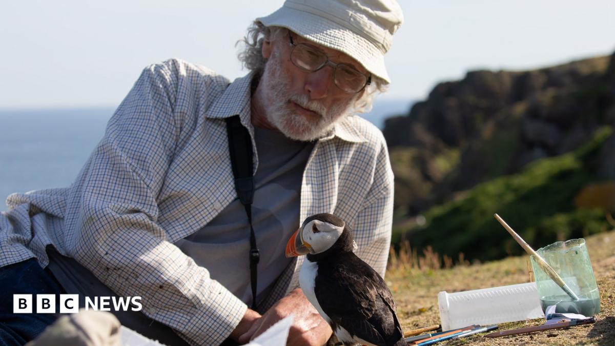 A man rests on his side on the ground as a small puffin bird sits in front of him. There is a cup of water with a paintbrush in it next to them. The man wears a cream bucket hat, white chequered shirt with a grey top underneath, and glasses.