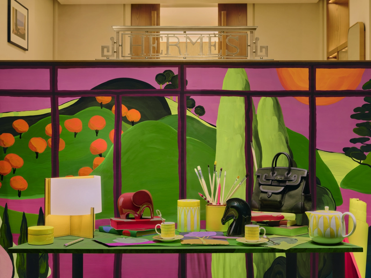 Colourful Hermès window display featuring Sarah Martinon's illustrated garden scene with vibrant pink, green and orange tones, styled with handbags, accessories and homeware products