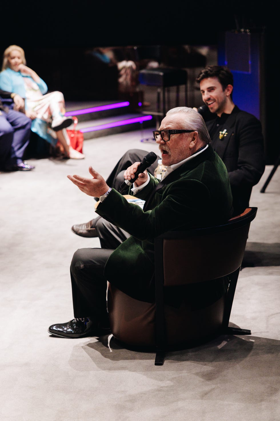 man in green jacket speaking into microphone during a group discussion.