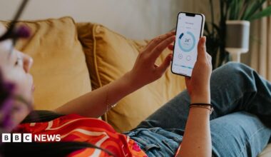 A picture of a woman in a red t shirt and jeans leaned back on a sofa checking her energy consumption on a mobile app.