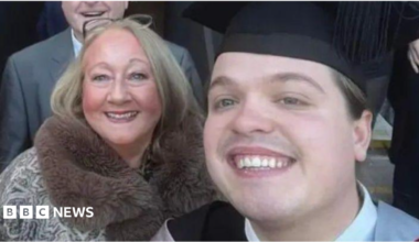 Image shows a middle-aged woman on the left, standing next to a young man in his graduation cap and gown. They are both smiling.