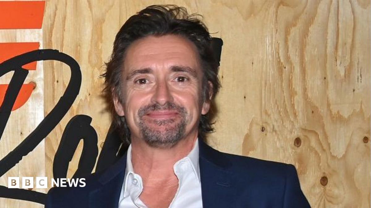 Richard Hammond is photographed from the shoulders up, standing in front of a promotional sign for the TV series 'The Grand Tour'. His face is facing slightly to the side and he is not looking at the camera. He is wearing a black suit jacket and a white shirt, with the top button undone.