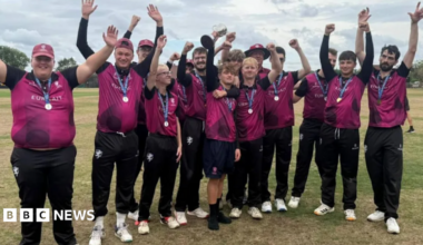 A team of young cricketers in purple shirts and black trousers all standing on the pitch with their hands waving in the air. They have small medals on ribbons round their necks.