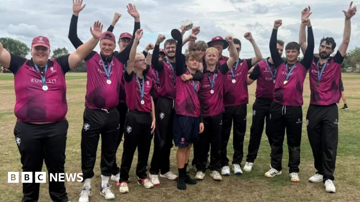 A team of young cricketers in purple shirts and black trousers all standing on the pitch with their hands waving in the air. They have small medals on ribbons round their necks.