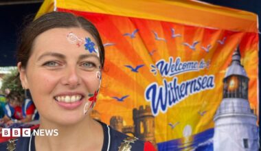 A woman is smiling at the camera. She has brown hair which has been tied up, and has red, white and blue flowers painted down the right-hand side of her face. She is standing in front of a flag which depicts a sunset and reads 'Welcome to Withernsea'.