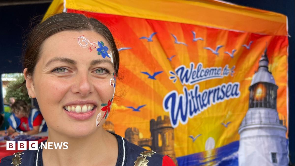 A woman is smiling at the camera. She has brown hair which has been tied up, and has red, white and blue flowers painted down the right-hand side of her face. She is standing in front of a flag which depicts a sunset and reads 'Welcome to Withernsea'.