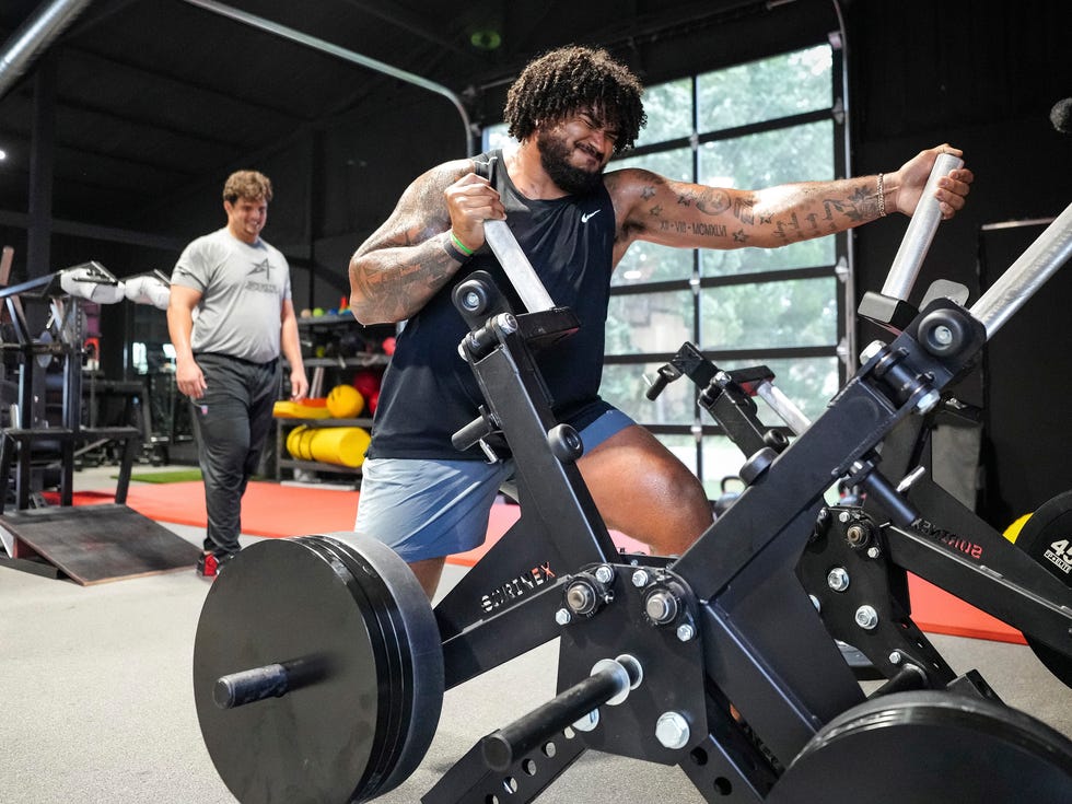 A person performs weight training on a machine in a gym while others observe. A person performs weight training on a machine in a gym while others observe.