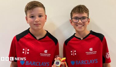 James and Harry are in their team kit on a red shirt with black trim and shoulder panels. It has the emblem of the MCC Foundation on the left hand side and a sponsors logo across the chest. They are holding their winners medal which has the MCC's coloured red and yellow ribbon on it.