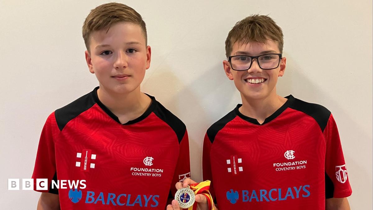 James and Harry are in their team kit on a red shirt with black trim and shoulder panels. It has the emblem of the MCC Foundation on the left hand side and a sponsors logo across the chest. They are holding their winners medal which has the MCC's coloured red and yellow ribbon on it.