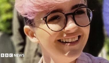 Imogen Nunn is smiling and looking off camera. She has pale pink hair and a pink checked jacket on, and is wearing dark rimmed round glasses.