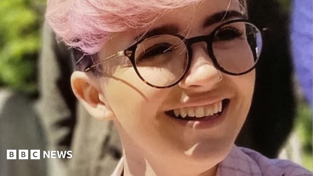 Imogen Nunn is smiling and looking off camera. She has pale pink hair and a pink checked jacket on, and is wearing dark rimmed round glasses.