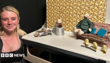 Stop-motion student Annie Buckeridge stands beside her small film set, which includes a fabric farmer dressed in a green coat and sitting on a tiny sofa next to three small yellow model chicks. Annie has long blonde hair, wears a dark top and is smiling at the camera.