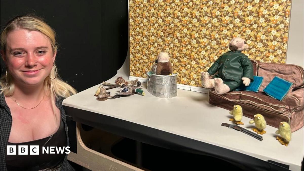 Stop-motion student Annie Buckeridge stands beside her small film set, which includes a fabric farmer dressed in a green coat and sitting on a tiny sofa next to three small yellow model chicks. Annie has long blonde hair, wears a dark top and is smiling at the camera.