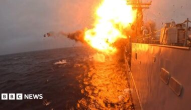 Orange flames and smokes erupts from an explosion on USS Klakring. Part of the grey warship is visible on the surface of a dark blue sea.