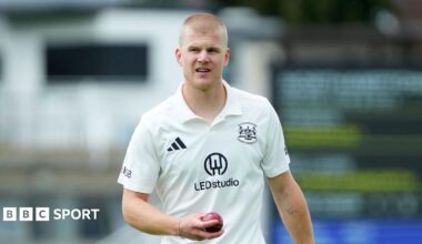 Luke Charlesworth holds a cricket ball in his right hand as he stands on the pitch. He is wearing white Gloucestershire kit