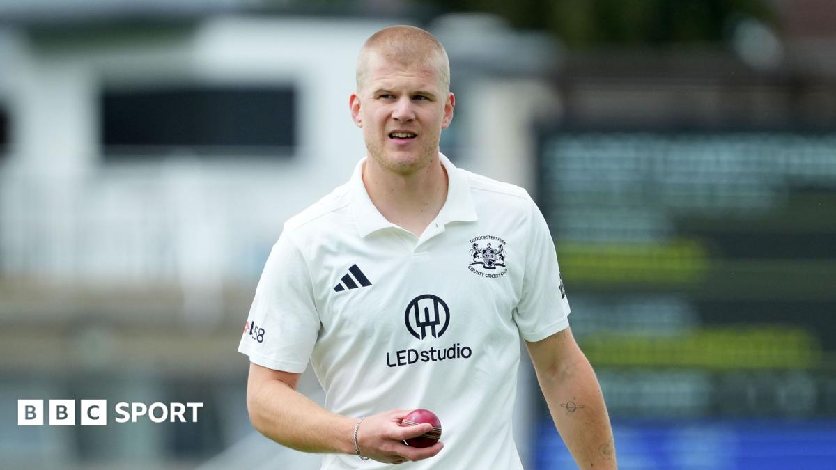 Luke Charlesworth holds a cricket ball in his right hand as he stands on the pitch. He is wearing white Gloucestershire kit