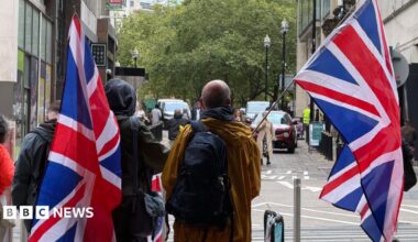 People carrying large Union Jack flags walk along a city street lined with office buildings and trees.