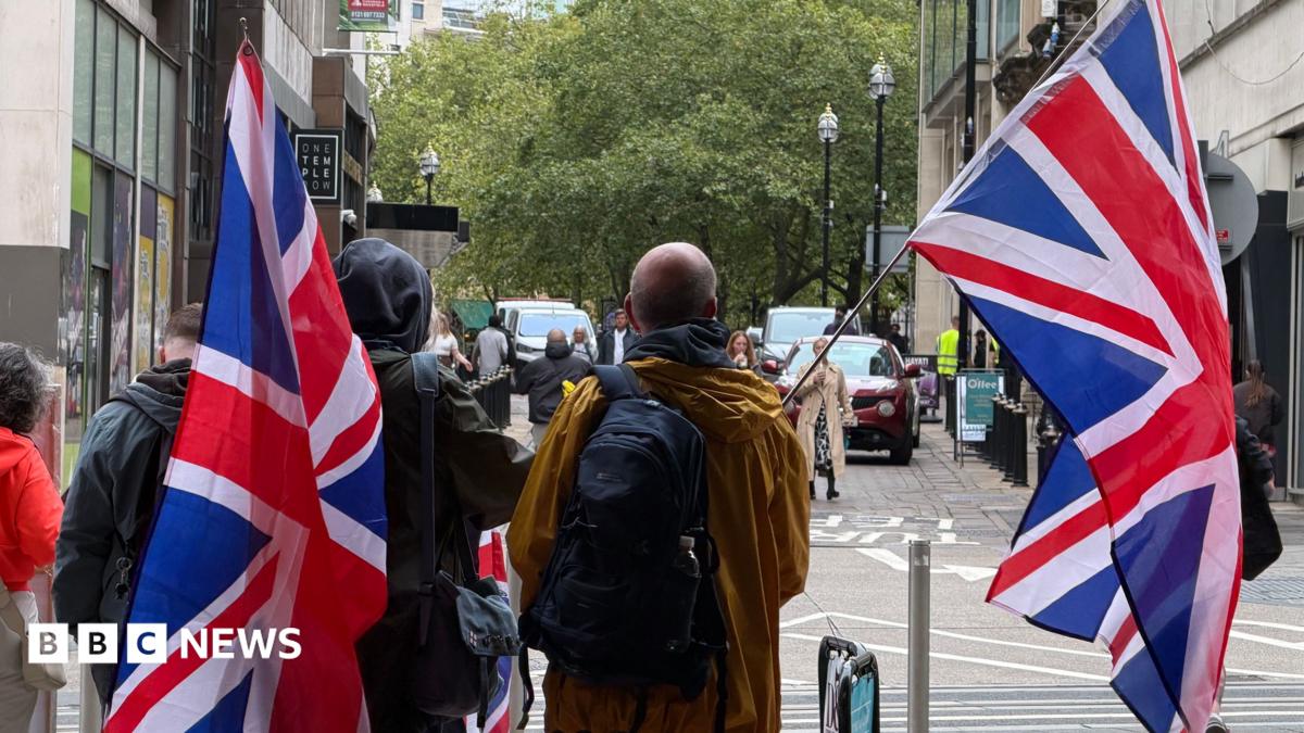 People carrying large Union Jack flags walk along a city street lined with office buildings and trees.