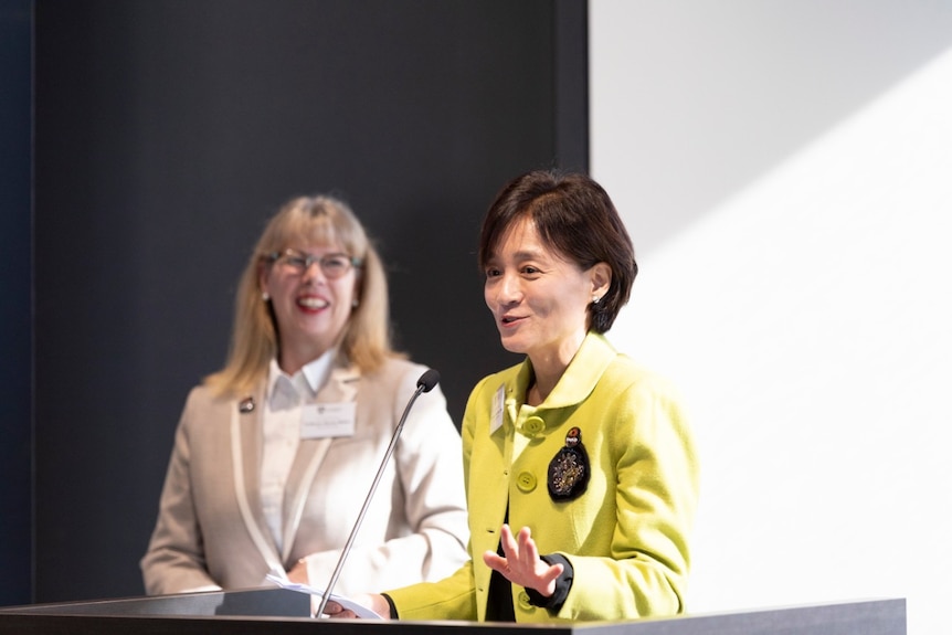 A woman in a green jacket speaks into a microphone at a lecture.