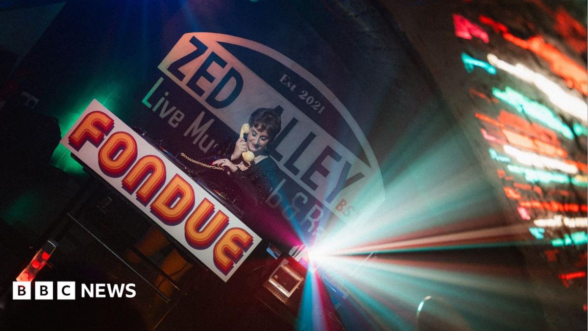 A woman holds a cream telephone to her ear as she stands behind a table with the word "fondue" printed on the front at the Zed Alley Live Music Bar.