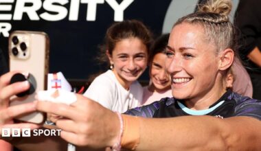 Natasha Hunt, wearing a blue England shirt, smiles while taking a selfie with a crowd of fans behind her
