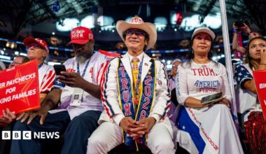 A man in a brick wall suit stands during the convention. He is holding a Make America Great Again hat. Beside him, a woman stands clapping