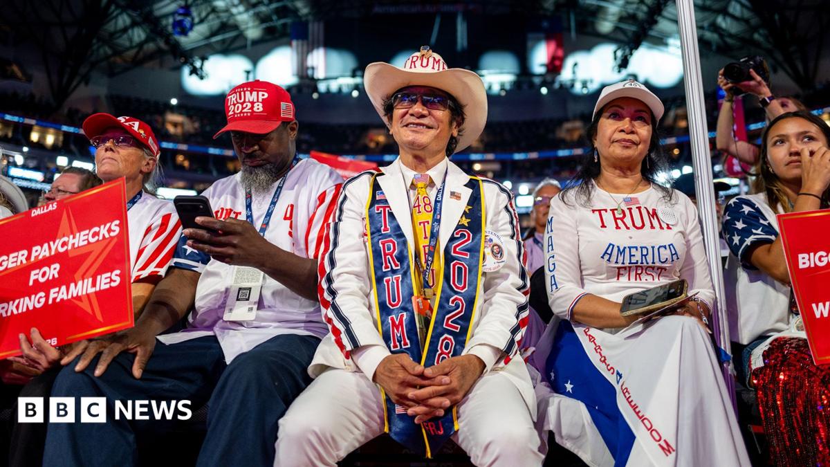 A man in a brick wall suit stands during the convention. He is holding a Make America Great Again hat. Beside him, a woman stands clapping