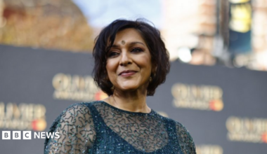 Meera Syal is looking at the camera, in front of a wall with brand names on it. She has her hair up and is wearing large gold earrings,