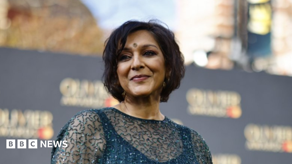 Meera Syal is looking at the camera, in front of a wall with brand names on it. She has her hair up and is wearing large gold earrings,