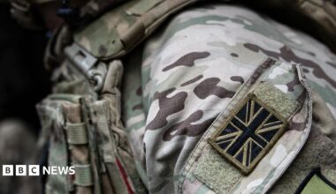 A close-up shot of a British soldier's uniform. A black and brown Union Jack patch is visible on their shoulder. The rest of the uniform is green, white and brown camouflage.