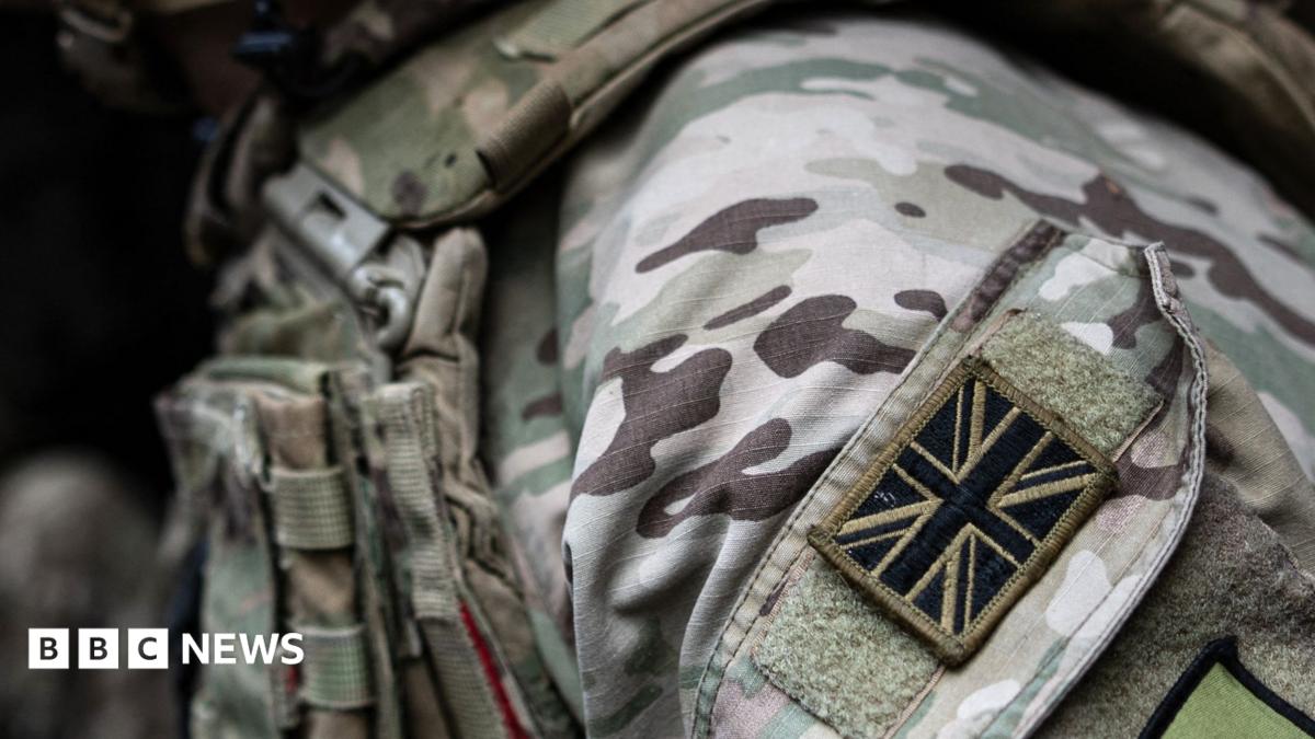 A close-up shot of a British soldier's uniform. A black and brown Union Jack patch is visible on their shoulder. The rest of the uniform is green, white and brown camouflage.