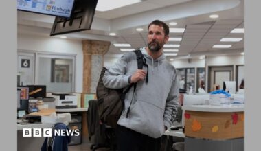 Dr. Michael “Robby” Robinavitch (Noah Wyle) looking exhausted, wearing black scrubs and holding a door open