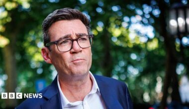 A medium close up of Andy Burnham wearing a blue suit and an open-collar white shirt with trees behind him in soft focus