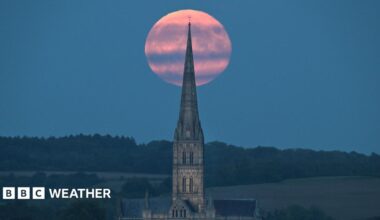 A pink-coloured full Harvest Moon rises behind Salisbury Cathedral spire. There are hills and trees in the distance and streaks of grey cloud across the Moon