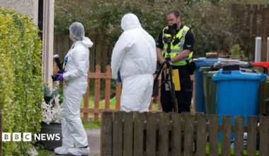 Two officers in white hazmat suits and head coverings in a garden.  One of them is carrying a camera. There is also a uniformed police officer in the garden and a camera on a tripod. There are also wheelie bins and fences.