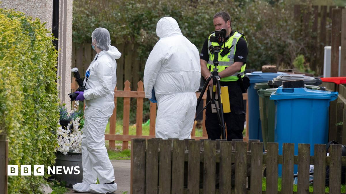 Two officers in white hazmat suits and head coverings in a garden.  One of them is carrying a camera. There is also a uniformed police officer in the garden and a camera on a tripod. There are also wheelie bins and fences.