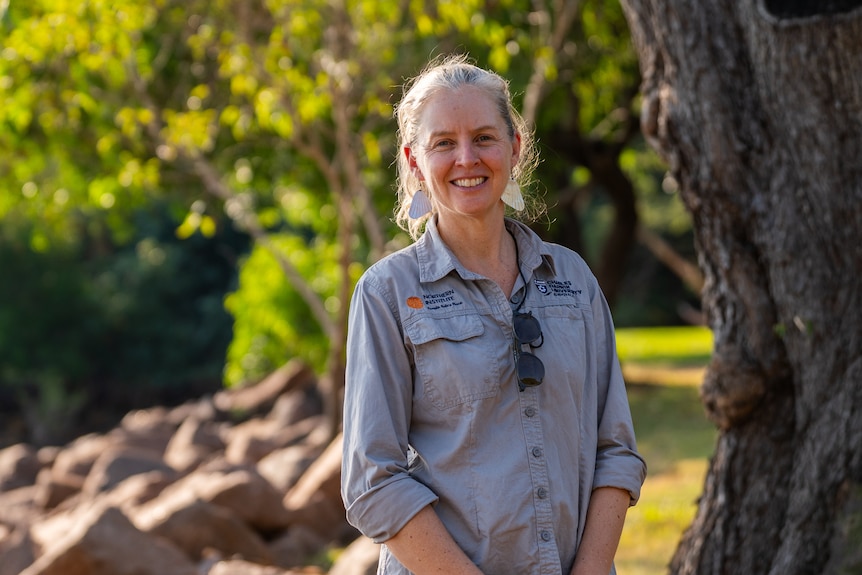 Rachel Groom, wearing a long-sleeved khaki shirt looks straight ahead smiling with trees in the background