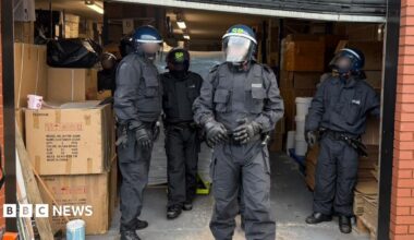 A group of four police officers in all-black uniforms and with helmets and masks are standing in a warehouse section of a red brick building.